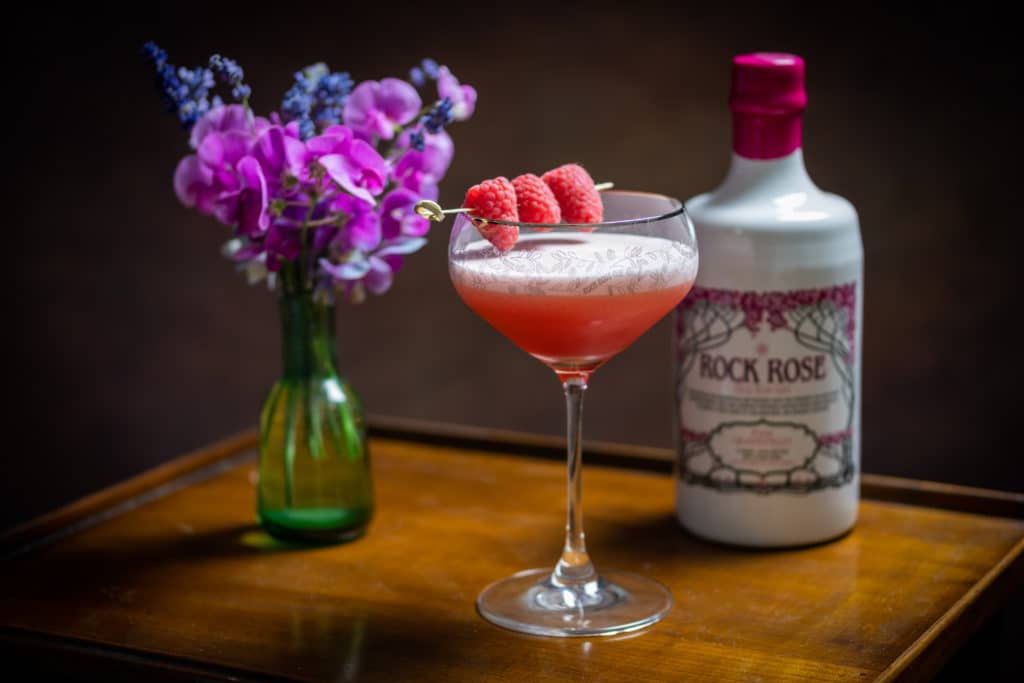 Bottle of Rock Rose Pink Grapefruit Old Tom Gin and Sour Old Tom cocktail served in a coupe glass and garnished with rasberries
