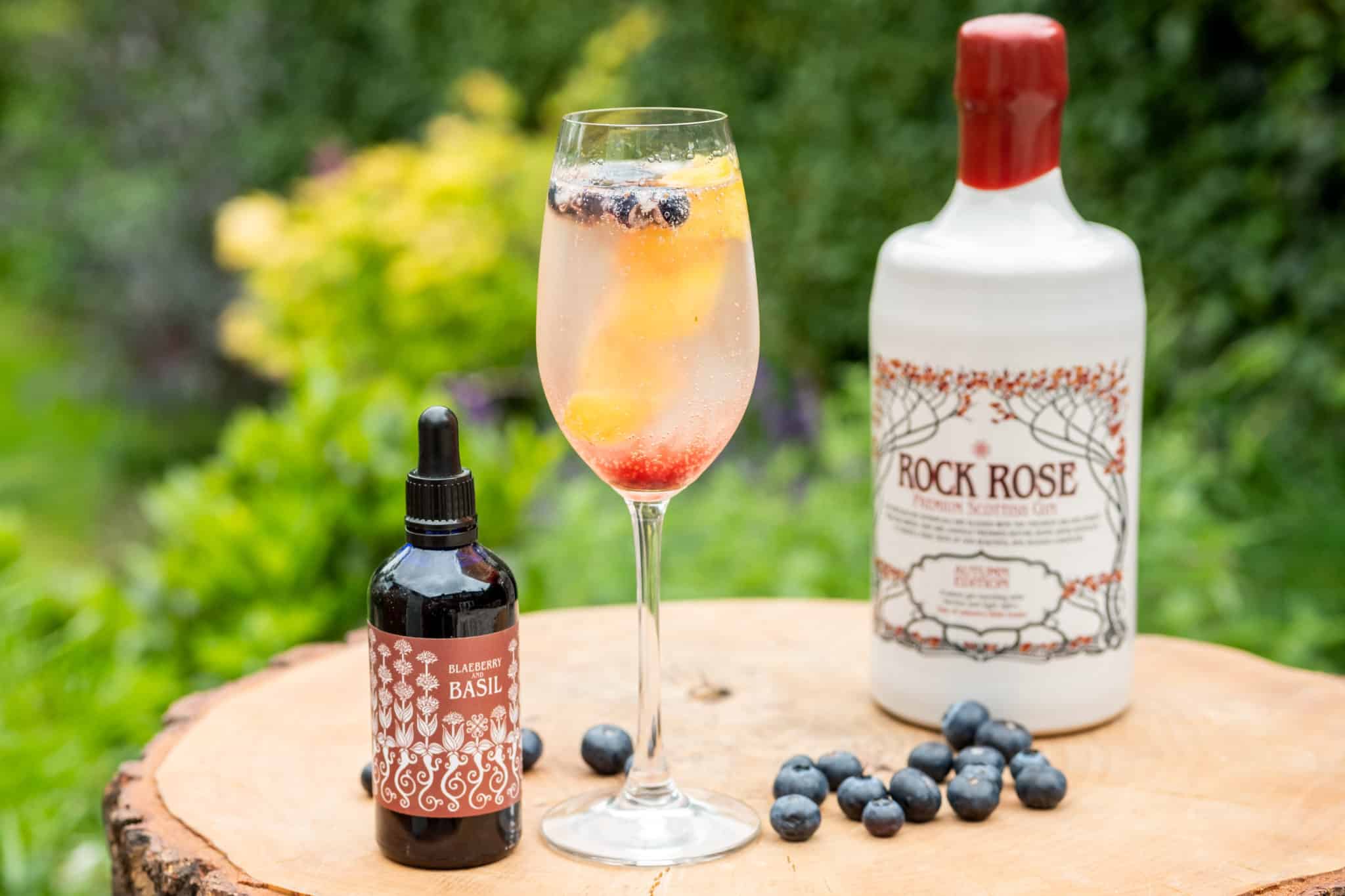Bottle of Rock Rose Gin Autumn Edition and bottle of Blaeberry and Basil liquid garnish with Highland 75 cocktail served in a flute glass and garnished with blueberries