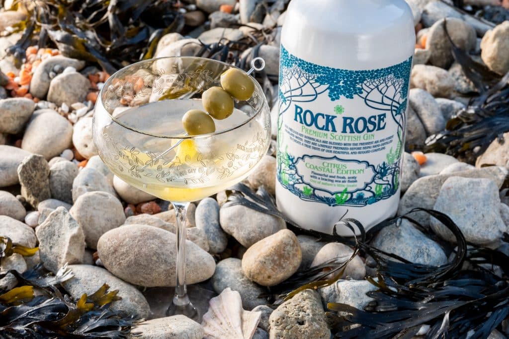 Bottle of Rock Rose Gin Citrus Coastal edition and Seashore Martini cocktail served in a coupe glass and garnished with lemon peel and olives