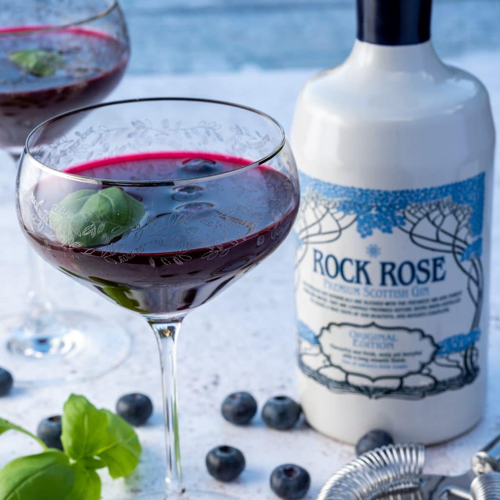 Bottle of Rock Rose Gin with Blueberry Basil Martini served in a coupe glass and garnished with blueberries and basil leaves