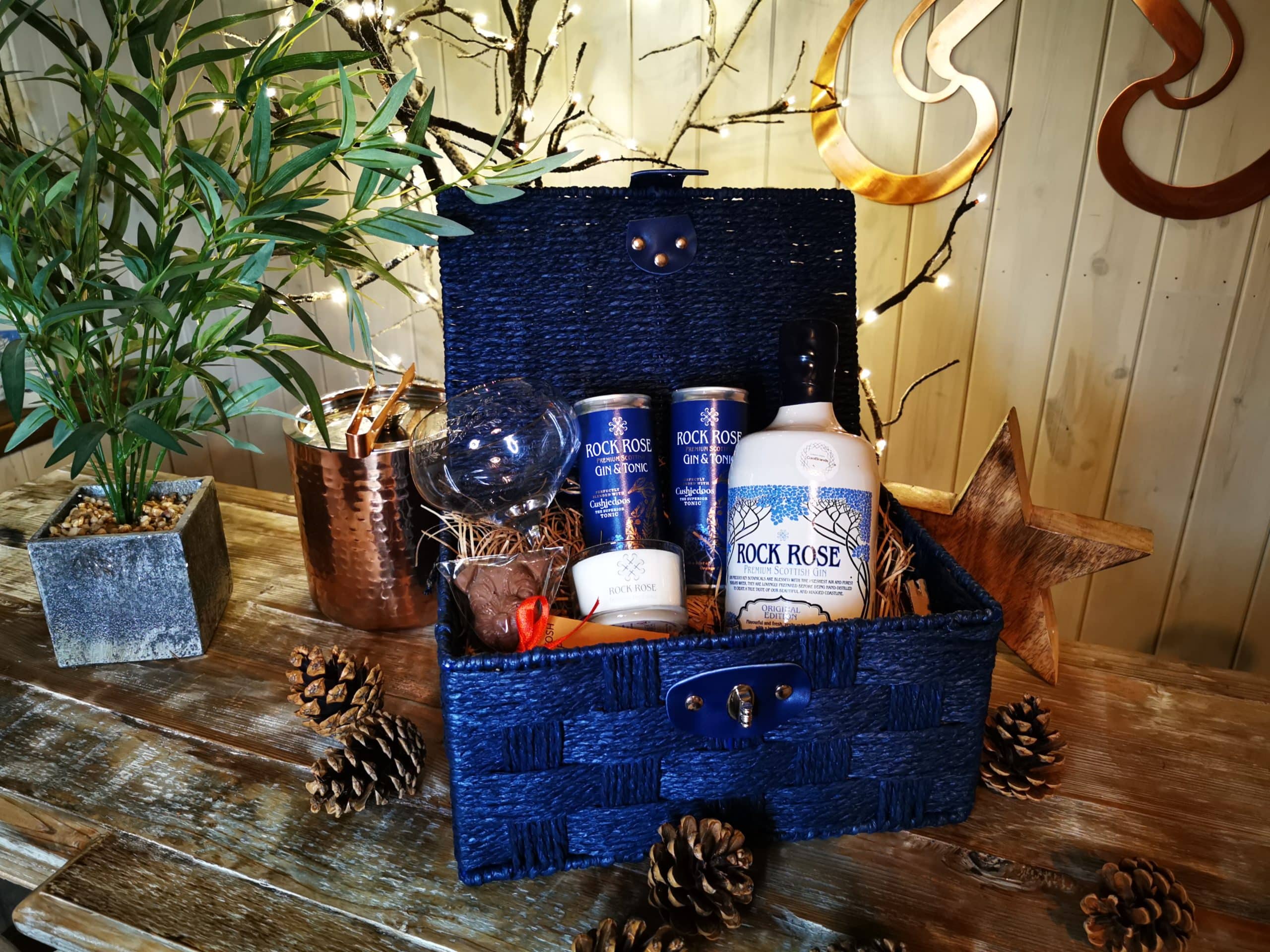 Content of the All Things Rock Rose Gin Hamper including a bottle of Rock Rose Gin, a branded glass, a small candle, 2 Rock Rose Gin & Cushiedoos Tonic cans and a Caithness Chocolate Mr Mackintosh Lollipup in a blue hamper