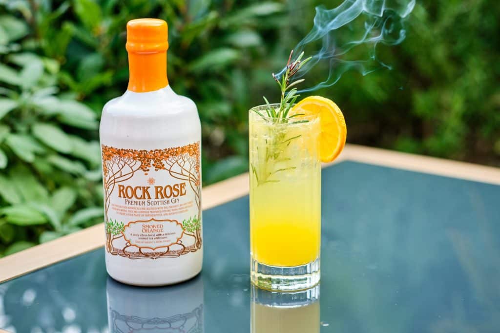 Lifestyle picture of a bottle of Rock Rose Gin Smoked Orange edition with Smoked Orange Spritz cocktail served in a tall glass with a slice of orange and a blow torched sprig of rosemary
